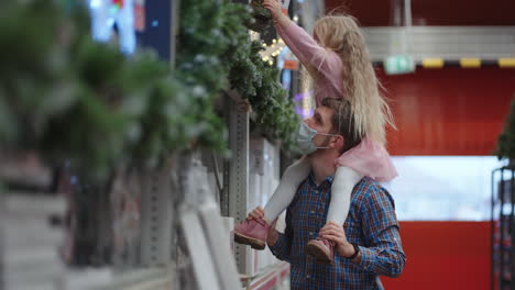 Father-and-daughter-sitting-on-their-shoulders-in-a-shopping-mall-choose-a-garland-for-the-house-and-Christmas-tree-for-Christmas-during-the-pandemic-in-slow-motion.